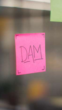 Vertical-Video-Close-Up-Of-Woman-Putting-Sticky-Note-With-DAM-Written-On-It-Onto-Transparent-Screen-In-Office